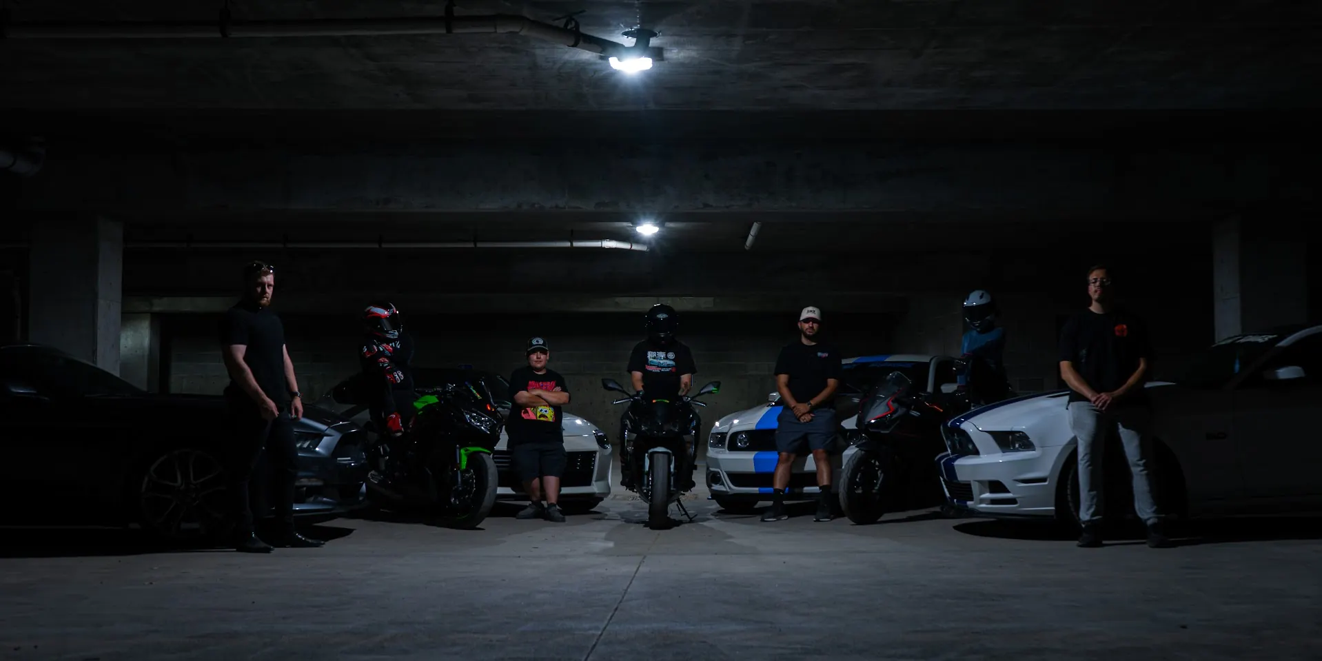 The CBB crew standing with their cars and bikes in a dark garage